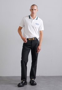 Vit polo t-shirt med grön randig krage, svarta byxor och kraftiga svarta skor. Bältet har guldtexten "MOSCHINO".