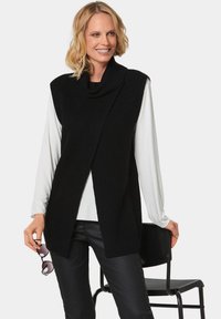 Schwarzer gestrickter ärmelloser Pullover mit einem Schlauchkragen und einem offenen Vorderdesign, getragen über einem weißen Langarmshirt, kombiniert mit schwarzen Hosen.