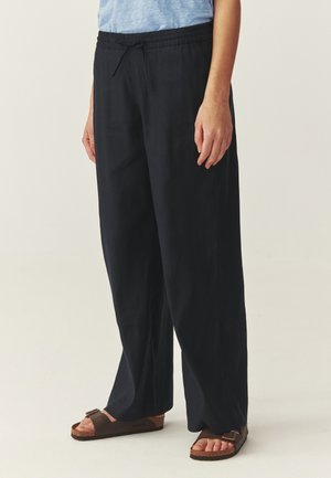 Osoba nosiaca voľné čierne nohavice s elastickým pásom a šnúrkou, skombinované s hnedými sandálmi s otvorenou špičkou, stojaca pred jednoliatym pozadím.