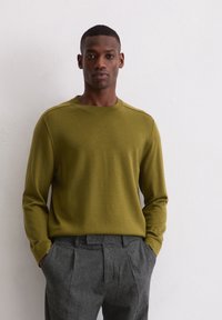 Man in een olijfgroene ronde hals trui en grijze geplooide broek, staand tegen een plain witte muur met zijn handen in de zakken.