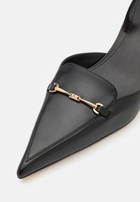 Chaussure noire en cuir pointue avec une embellissement doré sur le dessus. Texture lisse avec des détails de couture sur les côtés.