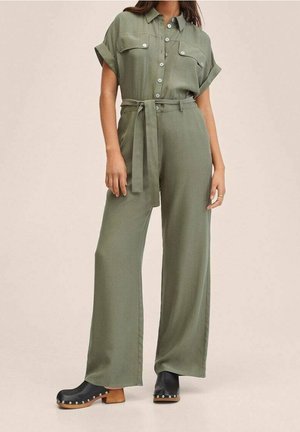 Woman wearing olive green short-sleeve jumpsuit with buttons, tie belt, wide legs, and black clogs with wooden heels and silver studs.