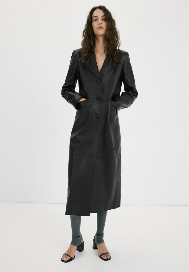 Femme aux longs cheveux bouclés portant un long manteau en cuir noir boutonné, des leggings gris et des sandales à talons noires, debout devant un fond blanc uni.