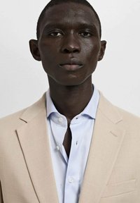 Beżowa marynarka na jasnoniebieskiej koszuli, z ostrym kołnierzykiem i jednym guzikiem. Gładka faktura z delikatnymi detalami przeszycia.