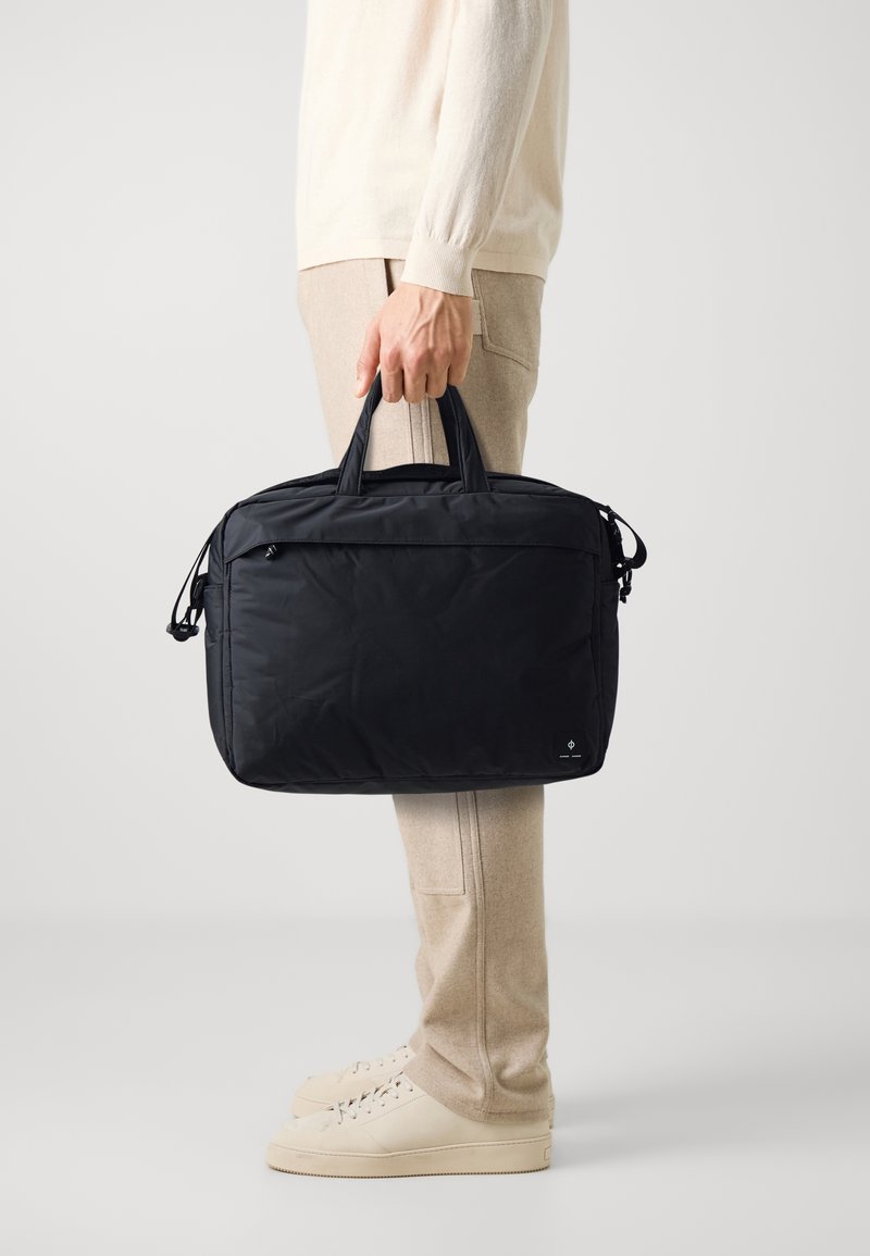 Sacoche d'ordinateur portable noire rembourrée avec doubles poignées supérieures, fermeture éclair et poches latérales. Tissu texturé et détail de marque subtil.