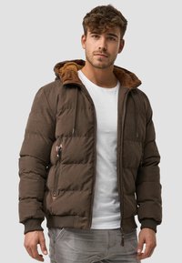 Brun polstret jakke med lynlås og hætte foret med tekstureret beige materiale. Har ribbede manchetter og to sidelommer.