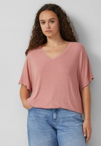 Roze geribbeld V-hals top met korte mouwen, met een relaxte pasvorm. Gecombineerd met lichtblauwe denim jeans, poseert het model met een hand in de zak.
