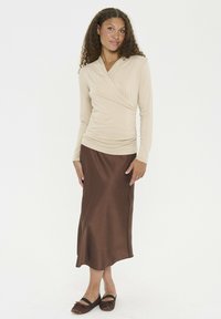 Kvinna med lockigt hår som bär en beige svepande långärmad topp, brun satin midi-kjol och bruna ballerina-skor, stående mot en vit bakgrund.