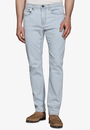 Hellblaue Jeans in enger Passform, getragen von einer stehenden Person, kombiniert mit braunen Wildleder-Loafern und einem in die Hose gesteckten weißen Hemd.