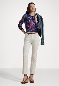 Μακρυμάνικη μπλούζα με λουλούδια σε μωβ και ροζ αποχρώσεις, λευκά τζιν με ίσια γραμμή, καφέ ζώνη και καφέ πλατφόρμες, με τζιν μπουφάν κρεμασμένο στον ώμο.