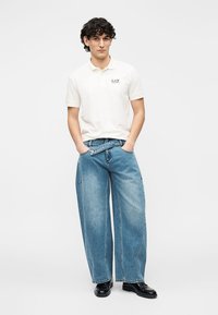 Λευκό πόλο με κοντά μανίκια και λογότυπο, μπλε τζιν παντελόνι με φαρδύ πόδι και μαύρα παπούτσια. Το μοντέλο στέκεται με τα χέρια στις τσέπες μπροστά σε απλό φόντο.