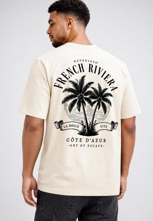 Mann von hinten gesehen, trägt ein beiges T-Shirt mit schwarzer Palmen-Grafik und Text "French Riviera, La Dolce Vita, Côte d'Azur, Art of Escape."