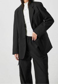 Zwarte op maat gemaakte blazer met gestructureerde schouders, een enkele knoopsluiting en een patchzak; gecombineerd met zwarte broeken met elastische tailleband.