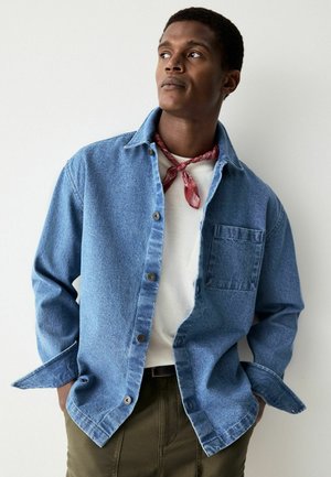 Hellblaues Denim-Hemd mit durchgehender Knopfleiste, Brusttasche und verlängerten Ärmeln; kombiniert mit einem weißen T-Shirt und einem roten Bandana.