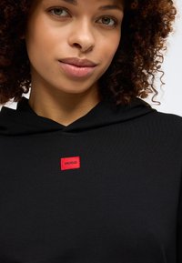 Fekete kapucnis pulóver, piros téglalap alakú címkével, rajta a "HUGO" logóval. Puha tapintású, lazán illeszkedő fazon, állítható kapucnival.