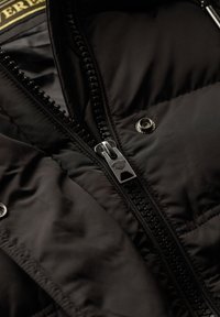 Chaqueta de plumas negra con una textura suave y acolchada, que presenta un cierre de cremallera plateada y botones a presión, destacando una construcción robusta.