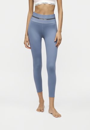 Persoon die blauwe hooggetailleerde Calvin Klein legging en een witte Calvin Klein sportbeha draagt, staat blootsvoets tegen een effen achtergrond.
