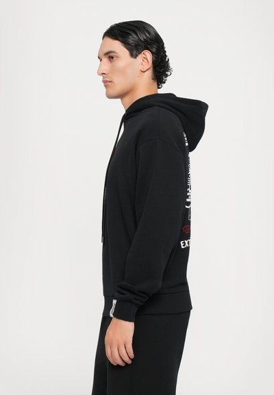 Sweatshirt à capuche noir avec cordons de serrage, arborant un texte imprimé sur le côté et une finition texturée. Le tissu semble doux et décontracté.