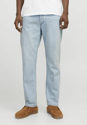 Man die lichtblauwe jeans met rechte pijpen draagt, een wit overhemd en bruine suède instapschoenen, staande tegen een effen achtergrond.