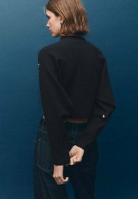 Maglione nero cropped con maniche lunghe e morbide, caratterizzato da un bottone decorativo sul polso. Indossato con jeans di denim scuri a vita alta.