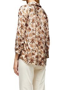 Blouse florale avec une texture soyeuse, présentant des motifs orange et marron sur un fond crème. Manches roulées et un ourlet arrondi.