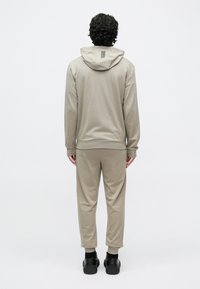 Göndör hajú férfi háttal áll, bézs kapucnis pulóvert és hozzáillő tréningnadrágot visel fekete sportcipővel fehér háttér előtt.