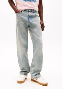 Jeans larges bleu clair avec un délavage, comprenant cinq poches et une fermeture éclair. Associés à des baskets blanches.