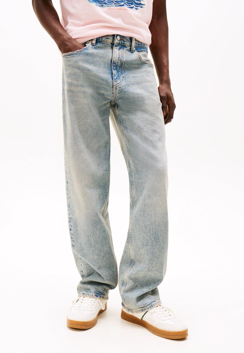 Jeans larges bleu clair avec un délavage, comprenant cinq poches et une fermeture éclair. Associés à des baskets blanches.