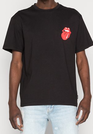Homme portant un t-shirt noir avec un petit logo rouge représentant une langue et des lèvres sur la poitrine gauche, associé à un jean bleu clair.