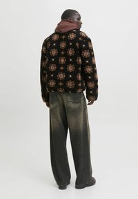 Giacca in fleece nera con motivi marroni e dorati, indossata sopra un hoodie bordeaux. Abbinata a pantaloni in denim sbiaditi a gamba larga.