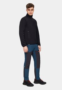 Czarna polarowa kurtka z wysokim kołnierzem i zamykanymi na zamek kieszeniami, zestawiona z niebieskimi i ciemnoszarymi spodniami w wielowarstwowym stylu oraz czarnymi butami do wędrówek.