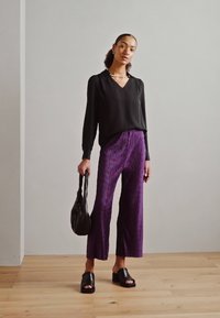 Blusa nera a maniche lunghe, scollo a V profondo, abbinata a pantaloni larghi plissettati viola e sandali neri con tacco a blocco. Porta una borsa nera.
