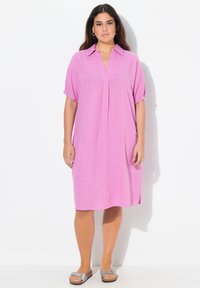Rosa linneklänning med korta upprullade ärmar, en v-ringning och sidoflikar. Matchad med silverfärgade sandaler på en vit bakgrund.
