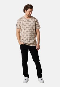Beige t-shirt featuring a palm tree pattern, short sleeves, and a crew neckline, paired with black jeans and white sneakers.