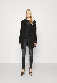 Zwarte lange jas met een gestructureerde kraag, gedragen over een donkere top. Gecombineerd met donkere jeans en schoenen met een patroon in leeuwenspot en geel.