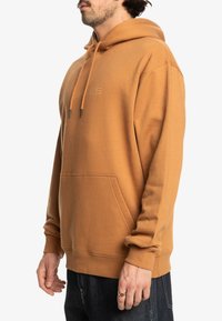 Tan hoodie gemaakt van een zachte, gestructureerde stof met een voorzak en koord. Beschikt over een subtiele logo in dezelfde kleur.