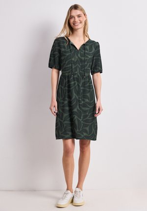 Kvinde smilende iført en mørkegrøn knælængde kjole med bladmotiver, korte ærmer og hvide sneakers, stående mod en ensfarvet baggrund.