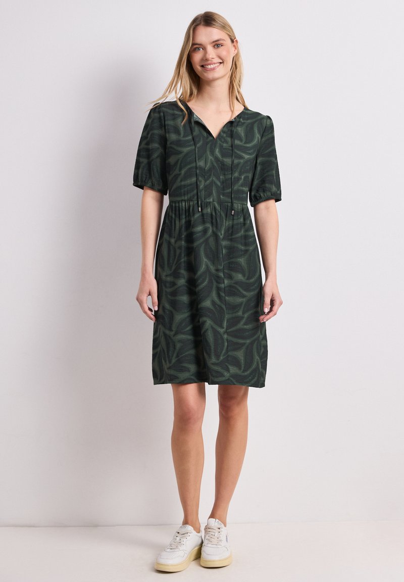 Femme souriante portant une robe vert foncé jusqu’aux genoux avec des motifs de feuilles, des manches courtes et des baskets blanches, debout devant un fond uni.