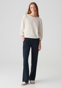 Weißer strukturierter Pullover mit Dreiviertelärmeln, kombiniert mit navyblauen weit geschnittenen Hosen und weißen Sneakers, vor einem neutralen Hintergrund stehend.