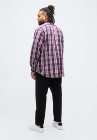 Άνδρας με ντρέντλοκς, που φοράει κόκκινο και μπλε πουκάμισο καρό, μαύρο παντελόνι και λευκά αθλητικά παπούτσια, στέκεται και κοιτάει πλάγια σε λευκό φόντο.