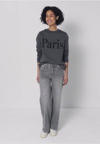 Grijze trui met "Paris" in grote zwarte letters, gecombineerd met lichtgrijze wijde jeans en witte sneakers. Gladde textuur, casual pasvorm.