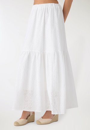 Gonna bianca alla caviglia con occhielli e balza arricciata, indossata con espadrillas con zeppa beige su sfondo chiaro.