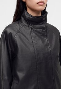 Femme portant une veste en cuir noire à col montant avec une fermeture à boutons au niveau du cou, montrant le haut du torse et une partie du visage.