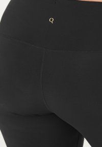 Zwarte leggings van rekbare stof, met een gouden "Q"-logo op de tailleband en een naadloos ontwerp voor een soepele pasvorm.