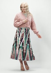 Różowy, dziany krótki sweter, srebrna bluzka z cekinami oraz wielokolorowa długa spódnica z cekinami w zielone, różowe i fioletowe wzory. Srebrne sandały.