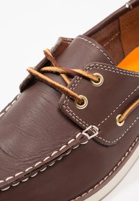 Braune Lederbootsschuhe mit gelb getönten Rohhautfäden, die durch Messing-Ösen gezogen sind, und weiße Nahtdetails.