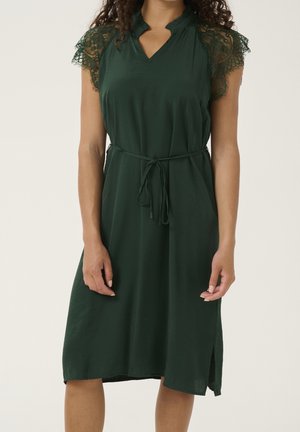 Mujer vestida con un vestido verde oscuro hasta la rodilla con mangas cortas de encaje y un cinturón fino en la cintura, de pie con las manos relajadas a los lados.