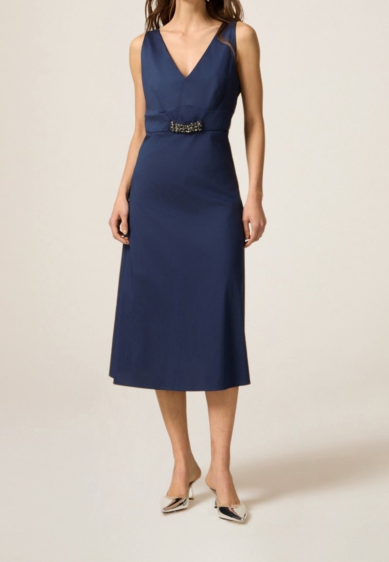 Femme portant une robe midi sans manches bleu marine à encolure en V avec une broche décorative à la taille, associée à des talons pointus argentés.