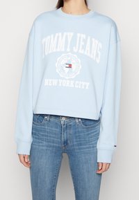 Persona che indossa un felpa leggera blu chiaro di Tommy Jeans con testo bianco del logo e jeans blu medio, in piedi contro uno sfondo semplice.