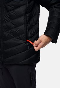 Giacca a vento nera con motivo a chevron, dotata di una tasca frontale con un'etichetta rossa etichettata "PACK AWAY." Mano che tiene il tirante della cerniera.